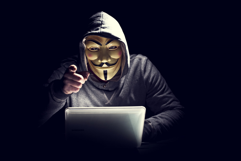 Person wearing a Guy Fawkes mask and hood with a laptop in front while pointing to forward.